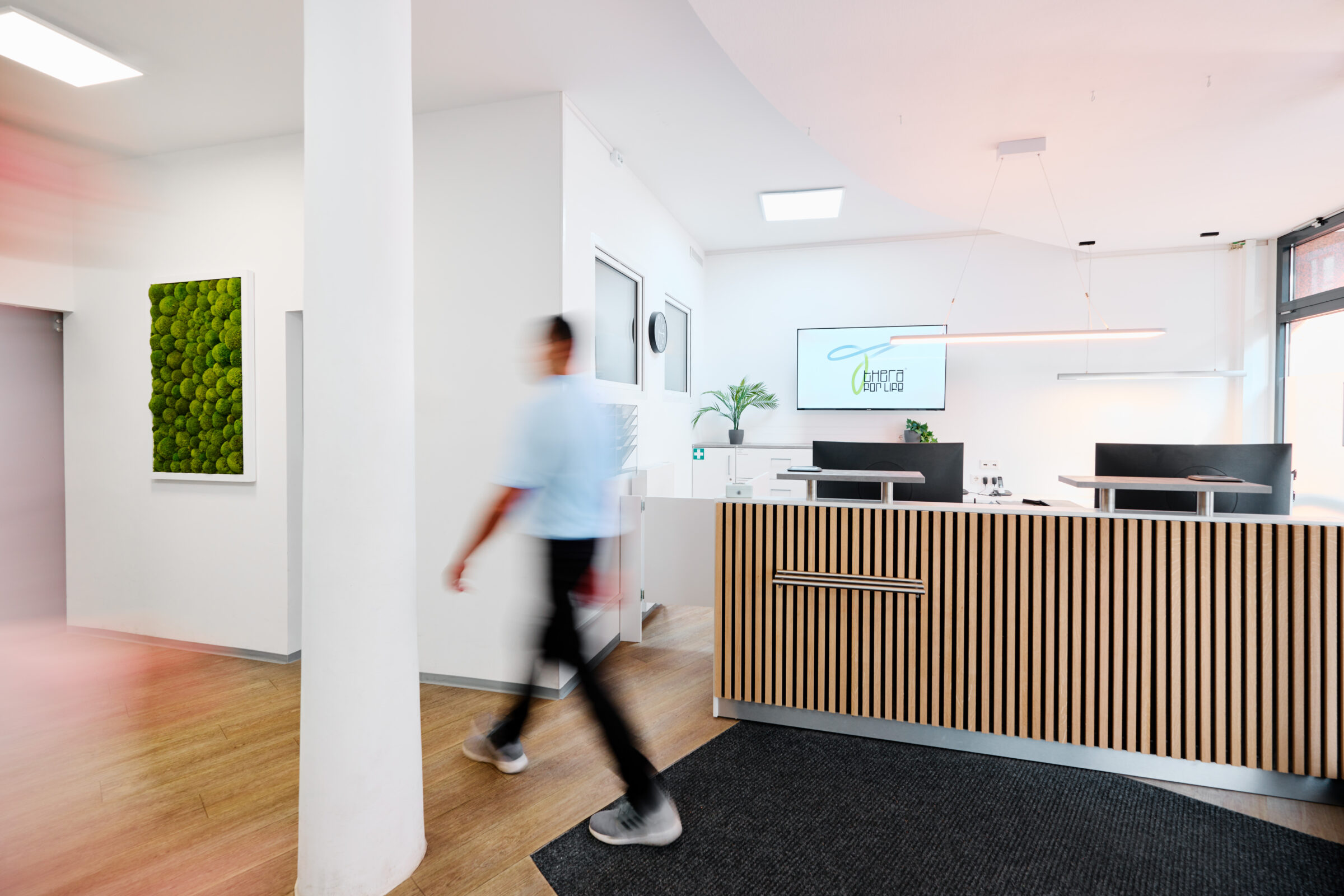 Moderner Empfangsbereich einer Praxis mit stilvollem Holzdesign, grünem Moosbild an der Wand und einem Monitor mit dem Logo "Thera for Life"; im Vordergrund läuft eine Person dynamisch durch den Raum.