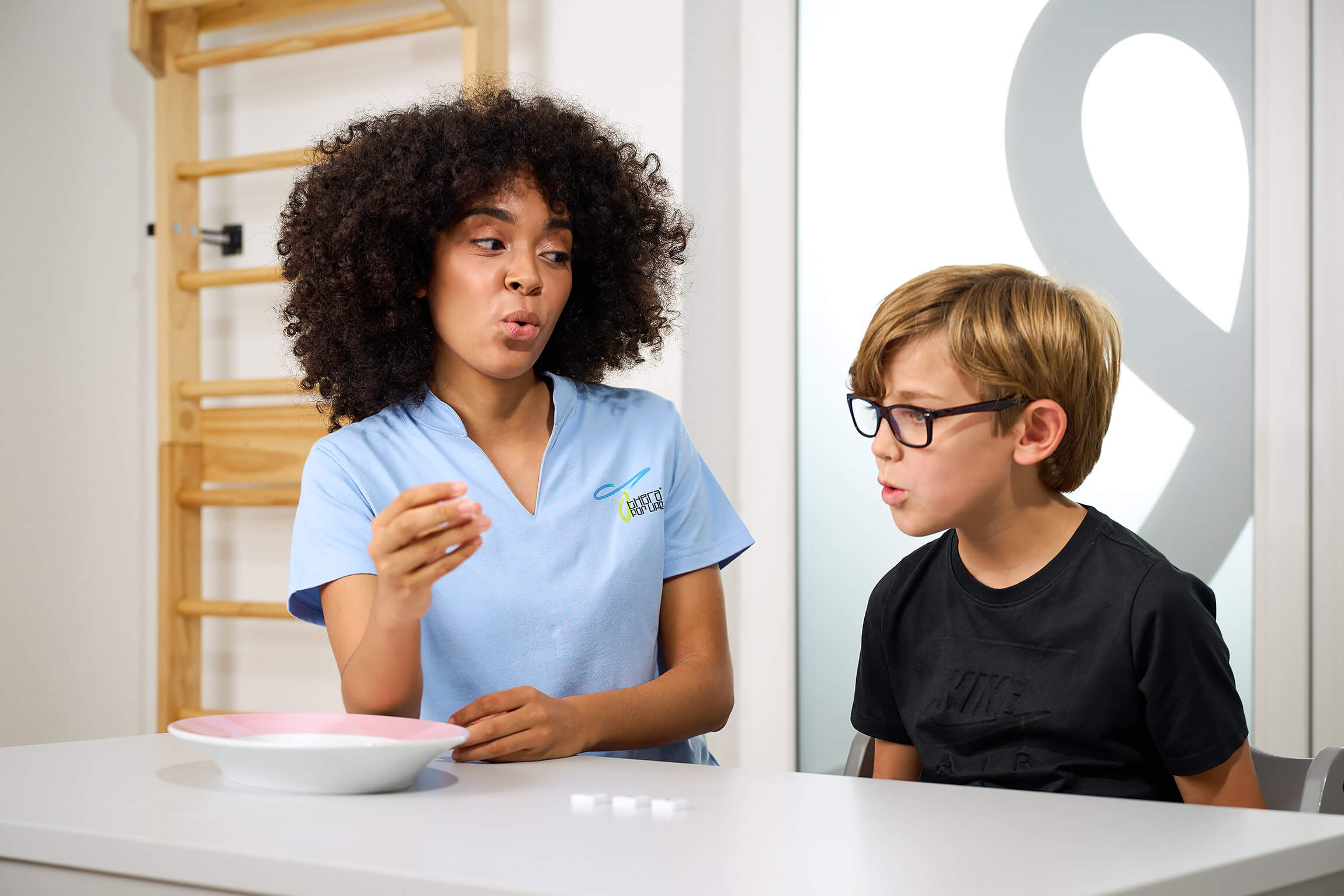 Therapeutin im blauen Thera for Life-Shirt arbeitet mit einem Jungen bei einer Sprachtherapiesitzung; beide sitzen an einem Tisch und üben artikulierte Laute.