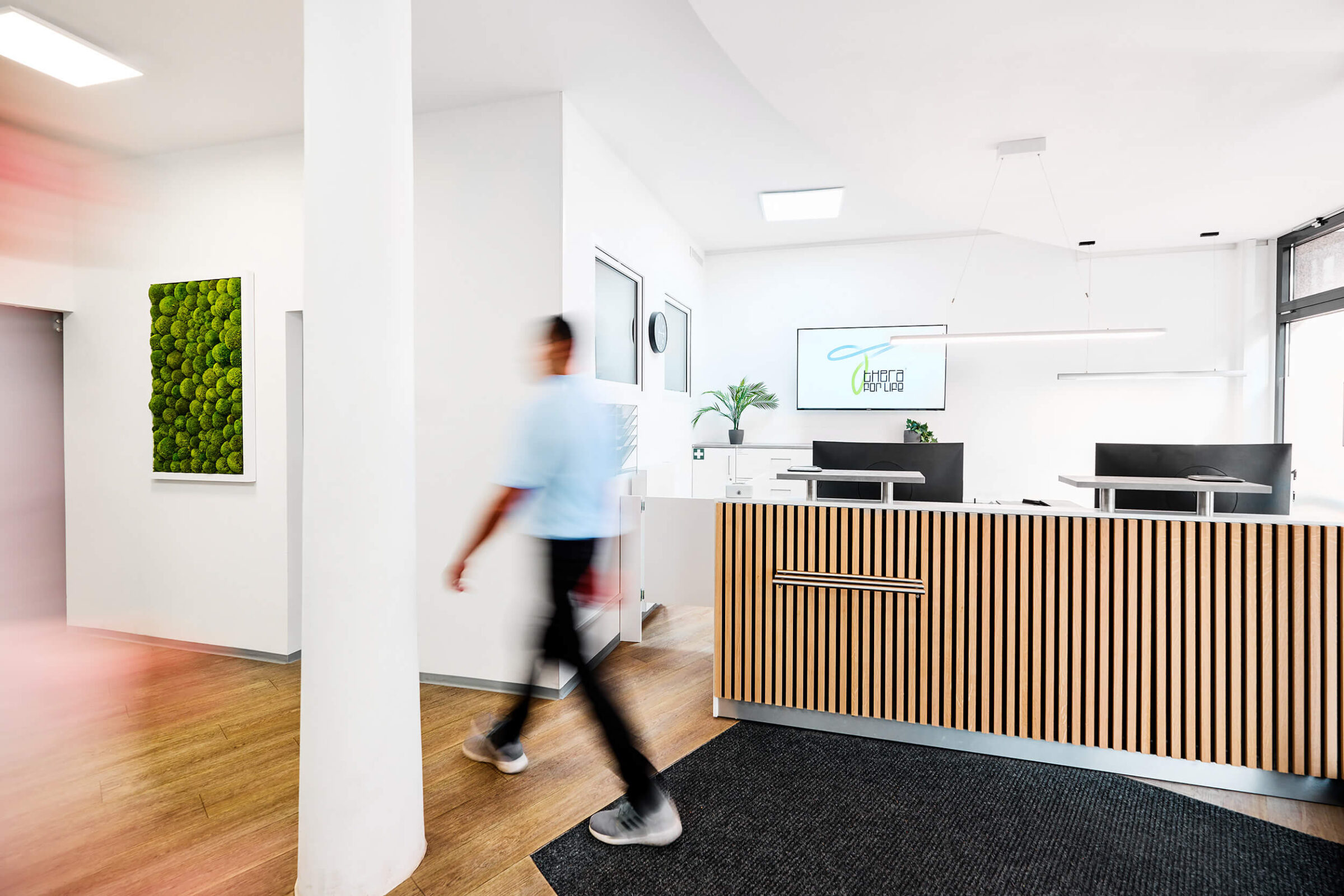 Moderner Empfangsbereich einer Praxis mit stilvollem Holzdesign, grünem Moosbild an der Wand und einem Monitor mit dem Logo "Thera for Life"; im Vordergrund läuft eine Person dynamisch durch den Raum.