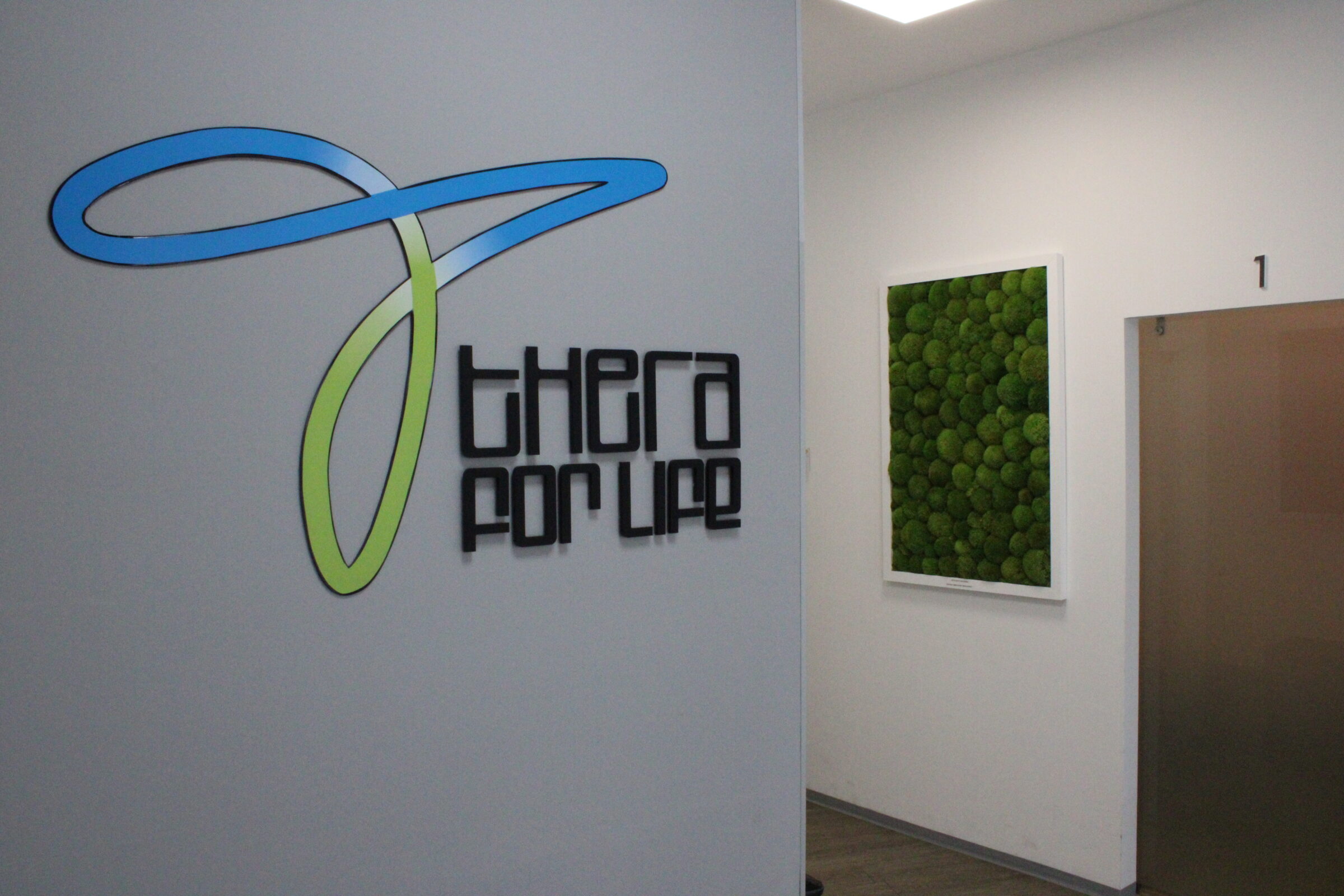 Innenansicht einer Praxis mit dem großen "Thera for Life"-Logo an der Wand, einer dekorativen grünen Mooswand und beschrifteten Türen zu den Behandlungsräumen.
