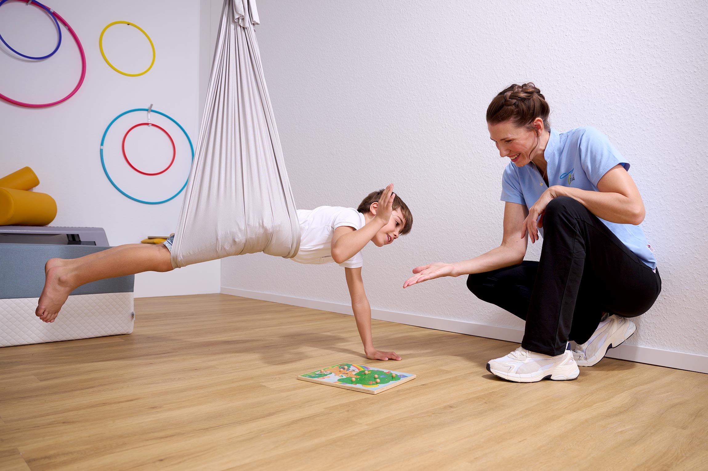 Ein Junge hängt in einem weißen Therapietuch, das an der Decke befestigt ist, und stützt sich mit einer Hand auf dem Boden ab. Vor ihm liegt ein Puzzle-Spiel. Eine Therapeutin in hellblauem Shirt und schwarzen Hosen kniet daneben und ermutigt ihn mit einer freundlichen Geste.