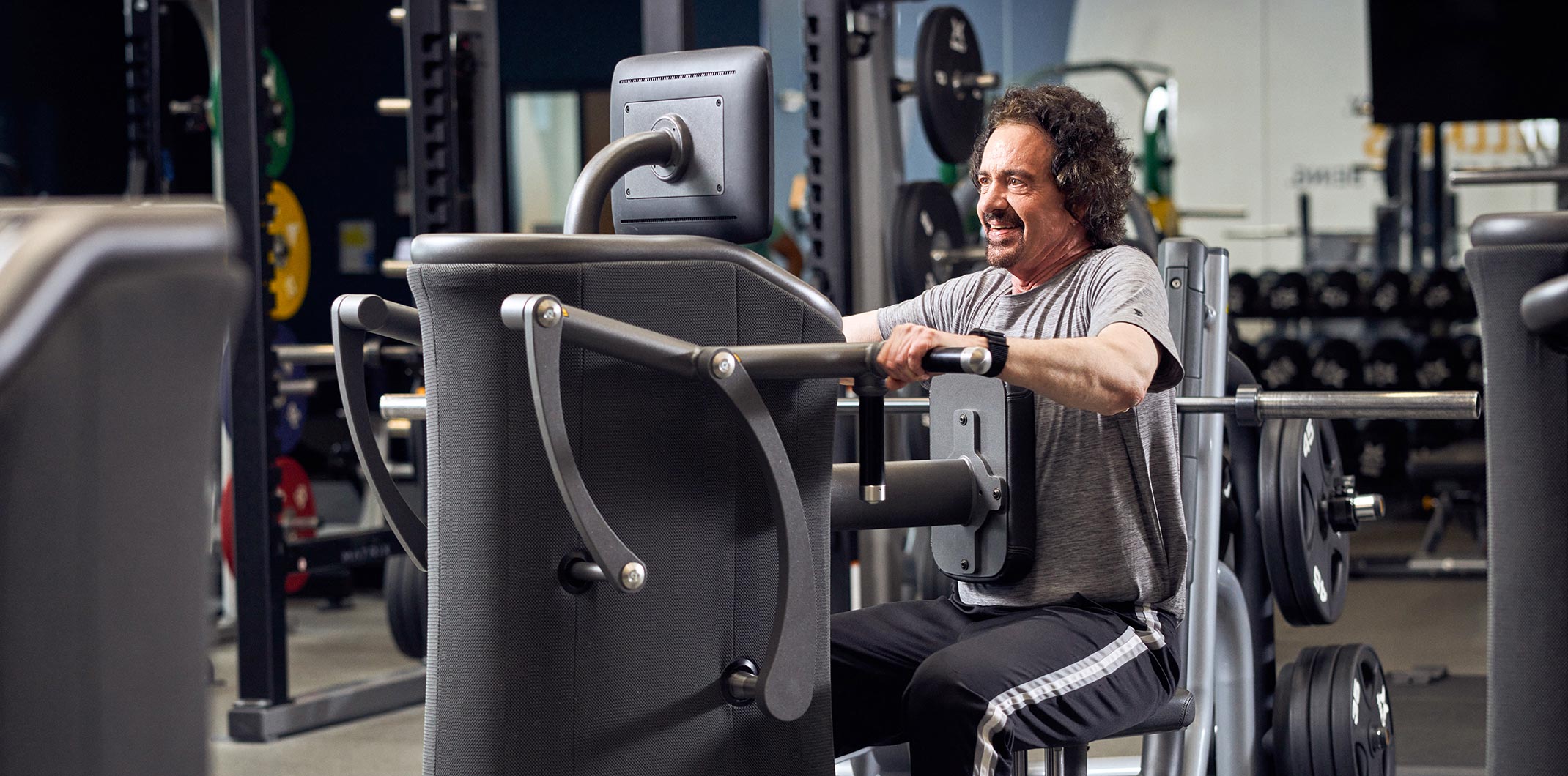 Ein Mann mittleren Alters trainiert an einer modernen Kraftmaschine in einem gut ausgestatteten Fitnessstudio. Im Hintergrund sind Hantelscheiben und weitere Trainingsgeräte sichtbar. Der Mann trägt ein graues T-Shirt, schwarze Trainingshose und wirkt konzentriert sowie motiviert.