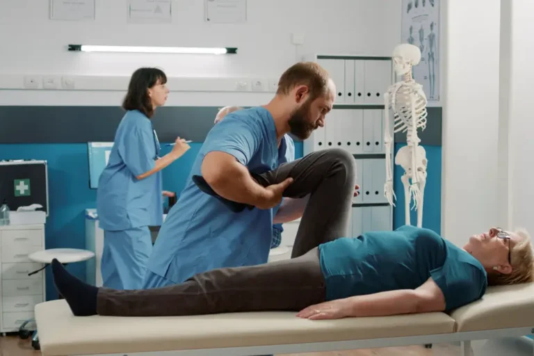 Ein Physiotherapeut in blauer Arbeitskleidung hilft einer älteren Frau bei einer Rehabilitationsübung. Die Frau liegt auf einer Behandlungsliege, während der Therapeut ihr Bein anhebt und bewegt. Im Hintergrund arbeiten weitere medizinische Fachkräfte, und ein menschliches Skelettmodell steht zur Veranschaulichung in der Ecke des Raumes.