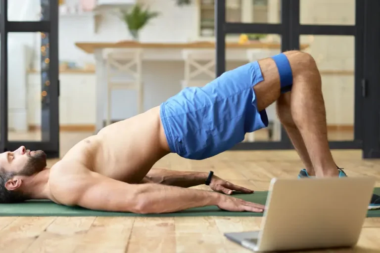 Ein athletischer Mann mit nacktem Oberkörper führt eine Brückenübung auf einer Yogamatte in einem gemütlichen Raum durch. Er verwendet ein Widerstandsband um die Oberschenkel und hat einen Laptop vor sich, was darauf hindeutet, dass er einem virtuellen Workout-Programm folgt. Die Szene vermittelt Fitness und Heimtraining.