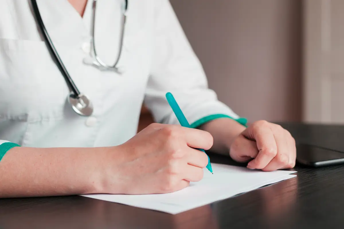 Nahaufnahme der Hände einer Ärztin, die mit einem Stift auf ein medizinisches Dokument schreibt. Ein Stethoskop ist um ihren Hals gehängt.