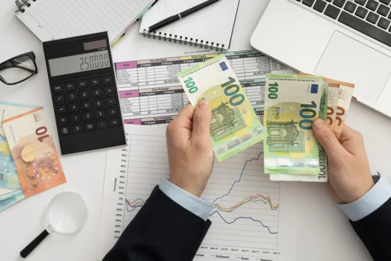 Flachlay-Aufnahme eines Arbeitsplatzes mit Finanzdokumenten, einem Laptop und einer Brille. Eine Person zählt Euro-Banknoten, während ein Taschenrechner mit einer eingegebenen Zahl sichtbar ist. Umgeben von Diagrammen und Notizbüchern symbolisiert das Bild finanzielle Planung und Buchhaltung.
