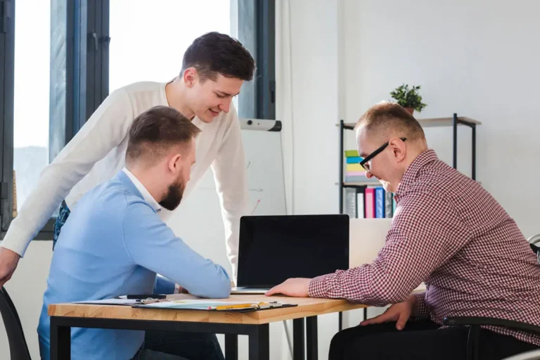 Drei Männer arbeiten gemeinsam in einem modernen Büro an einem Tisch. Sie sind in eine Diskussion vertieft und betrachten konzentriert den Bildschirm eines Laptops. Die Szene vermittelt Teamarbeit, Zusammenarbeit und eine produktive Atmosphäre.