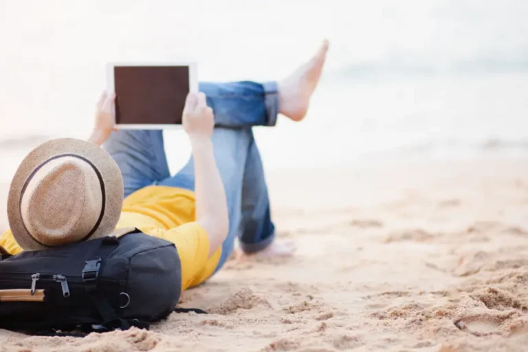 Eine Person liegt entspannt am Strand, trägt einen Strohhut und hält ein Tablet in der Hand. Neben ihr liegt ein schwarzer Rucksack. Im Hintergrund ist das Meer zu sehen, das eine ruhige und entspannte Atmosphäre vermittelt.
