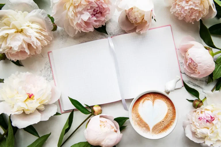 Ein stilvolles Arrangement aus zarten Pfingstrosen, einem offenen Notizbuch mit weißem Band und einer Tasse Kaffee mit Herz-Milchschaum. Die helle, weiche Ästhetik vermittelt eine ruhige und inspirierende Atmosphäre, ideal für kreative Gedanken oder Entspannung.
