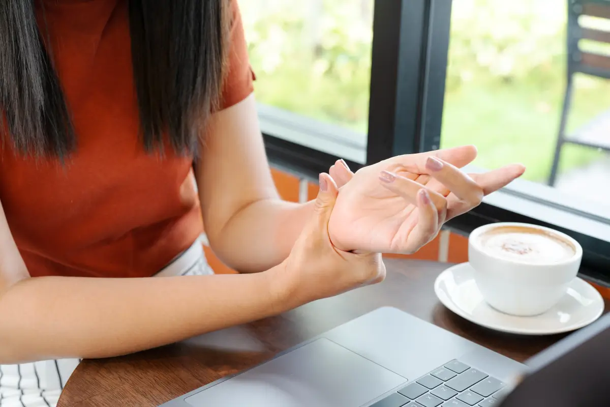 Eine Frau sitzt in einem Café vor einem Laptop und hält ihr Handgelenk mit der anderen Hand, als ob sie Schmerzen oder Unbehagen verspürt. Neben ihr steht eine Tasse Kaffee.