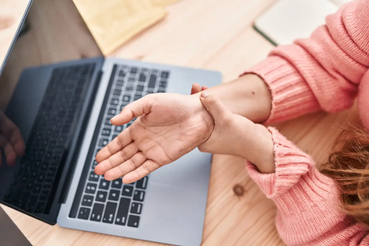 Eine junge Frau in einem rosa Pullover sitzt an einem Schreibtisch mit einem Laptop und hält ihr schmerzendes Handgelenk, möglicherweise aufgrund langer Arbeit am Computer.