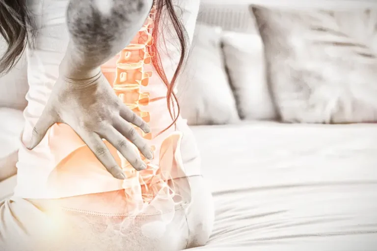 Frau sitzt auf einem Bett und hält sich den schmerzenden unteren Rücken; hervorgehobene Wirbelsäule symbolisiert Schmerzen.