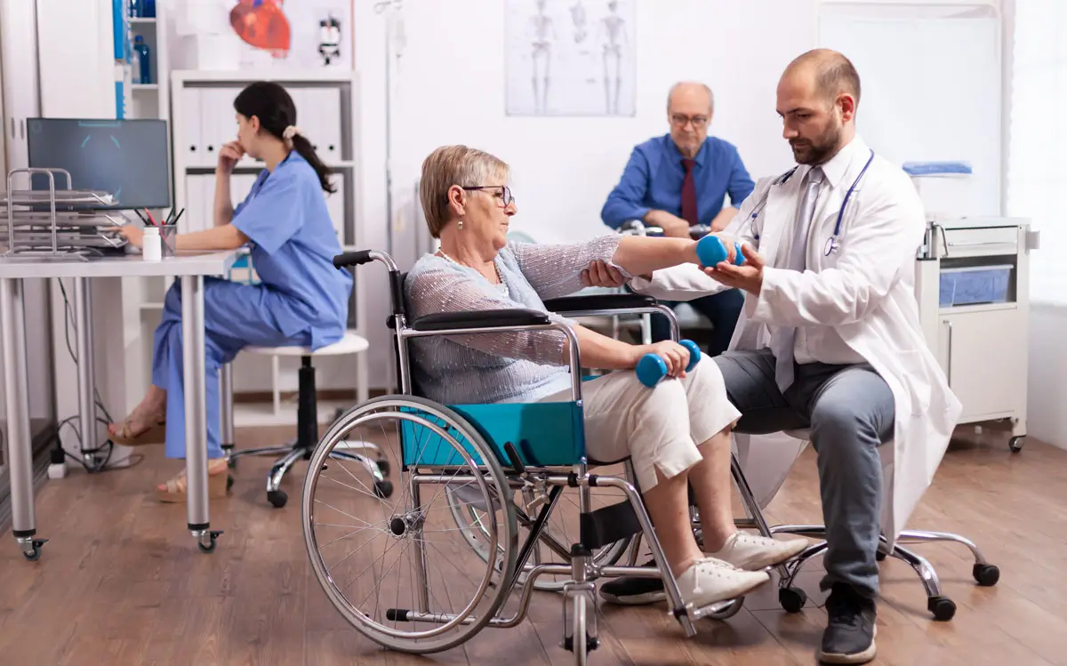 Ältere Frau im Rollstuhl trainiert mit Hanteln unter Anleitung eines Arztes in einer Reha-Klinik, während im Hintergrund weitere Patienten und medizinisches Personal arbeiten.
