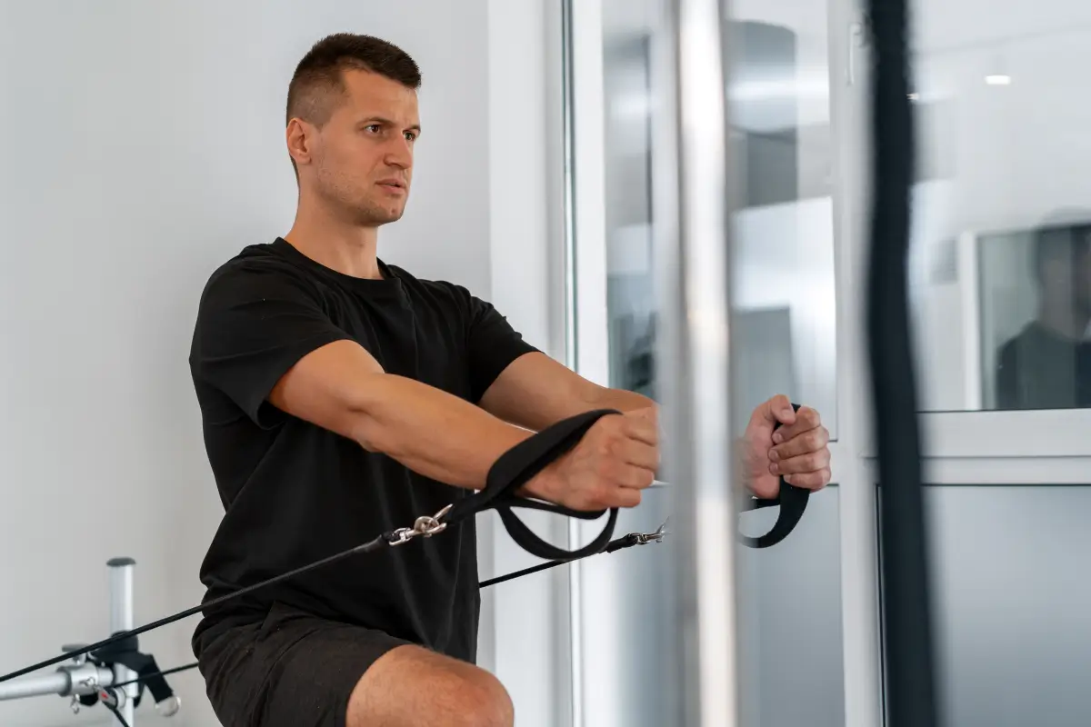Ein junger Mann mit kurzen Haaren und einem schwarzen T-Shirt trainiert an einem Seilzuggerät in einem modernen Fitnessstudio. Er sitzt auf einer Bank, hält die Griffe mit beiden Händen und konzentriert sich auf die Übung.