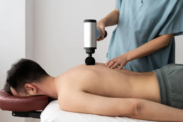 Ein Physiotherapeut verwendet eine Massagepistole auf dem Rücken eines liegenden Patienten zur Muskelentspannung.