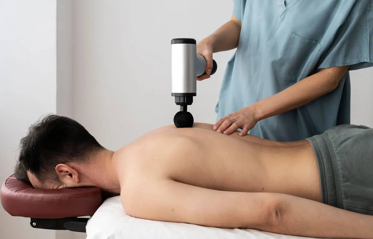 Ein Physiotherapeut verwendet eine Massagepistole auf dem Rücken eines liegenden Patienten zur Muskelentspannung.