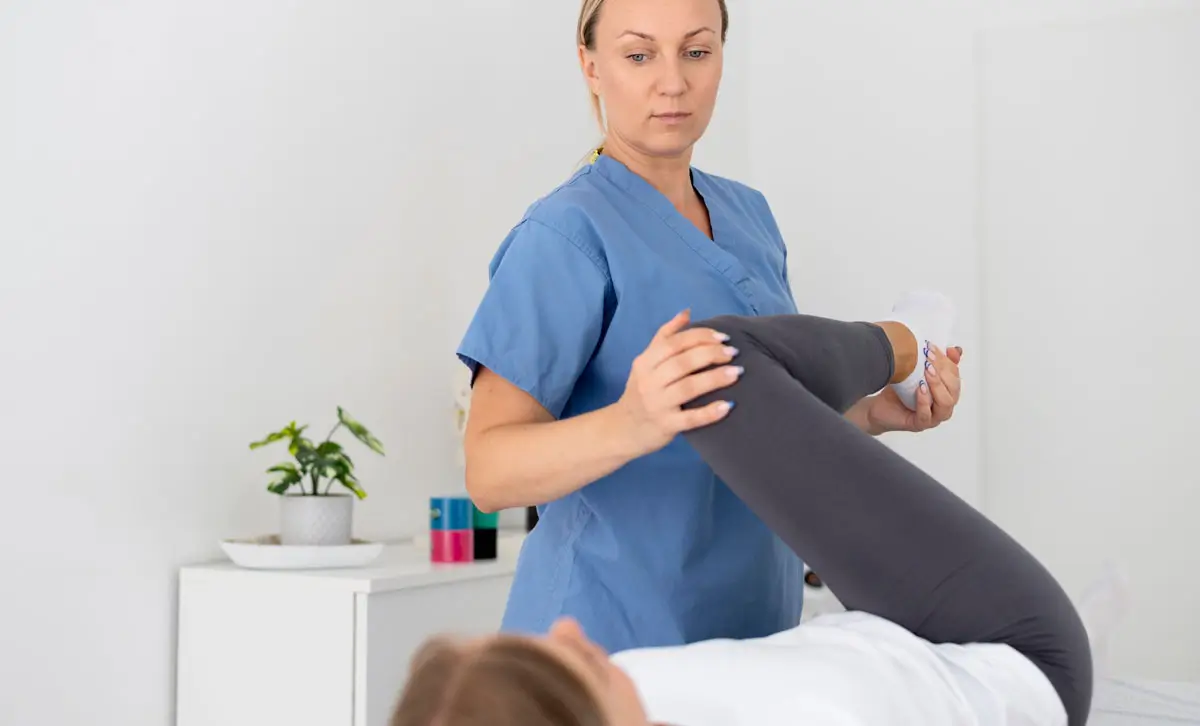 Physiotherapeutin in blauer Arbeitskleidung führt eine Beinmobilisation bei einer liegenden Patientin in einer modernen Praxis durch.