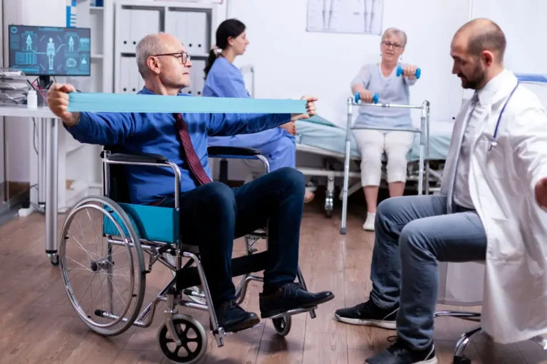 Älterer Mann im Rollstuhl macht physiotherapeutische Übungen mit einem Widerstandsband, während ein Arzt die Bewegung anleitet. Im Hintergrund trainieren weitere Patienten in einer Reha-Klinik