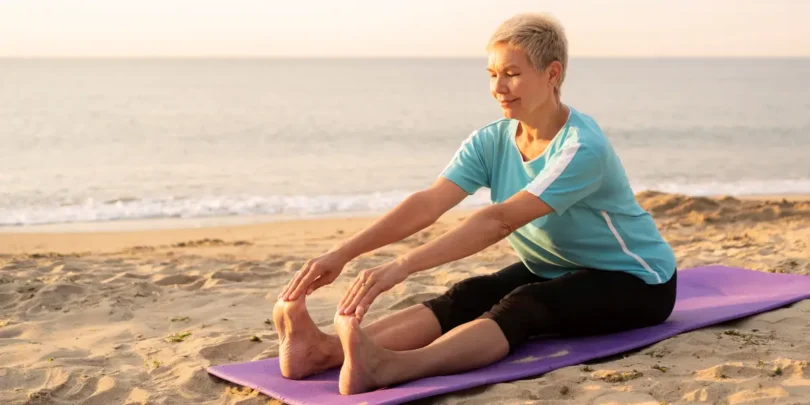 Seniorin in Sportkleidung sitzt auf einer Yogamatte am Strand, streckt sich und genießt die ruhige Atmosphäre am Wasser.