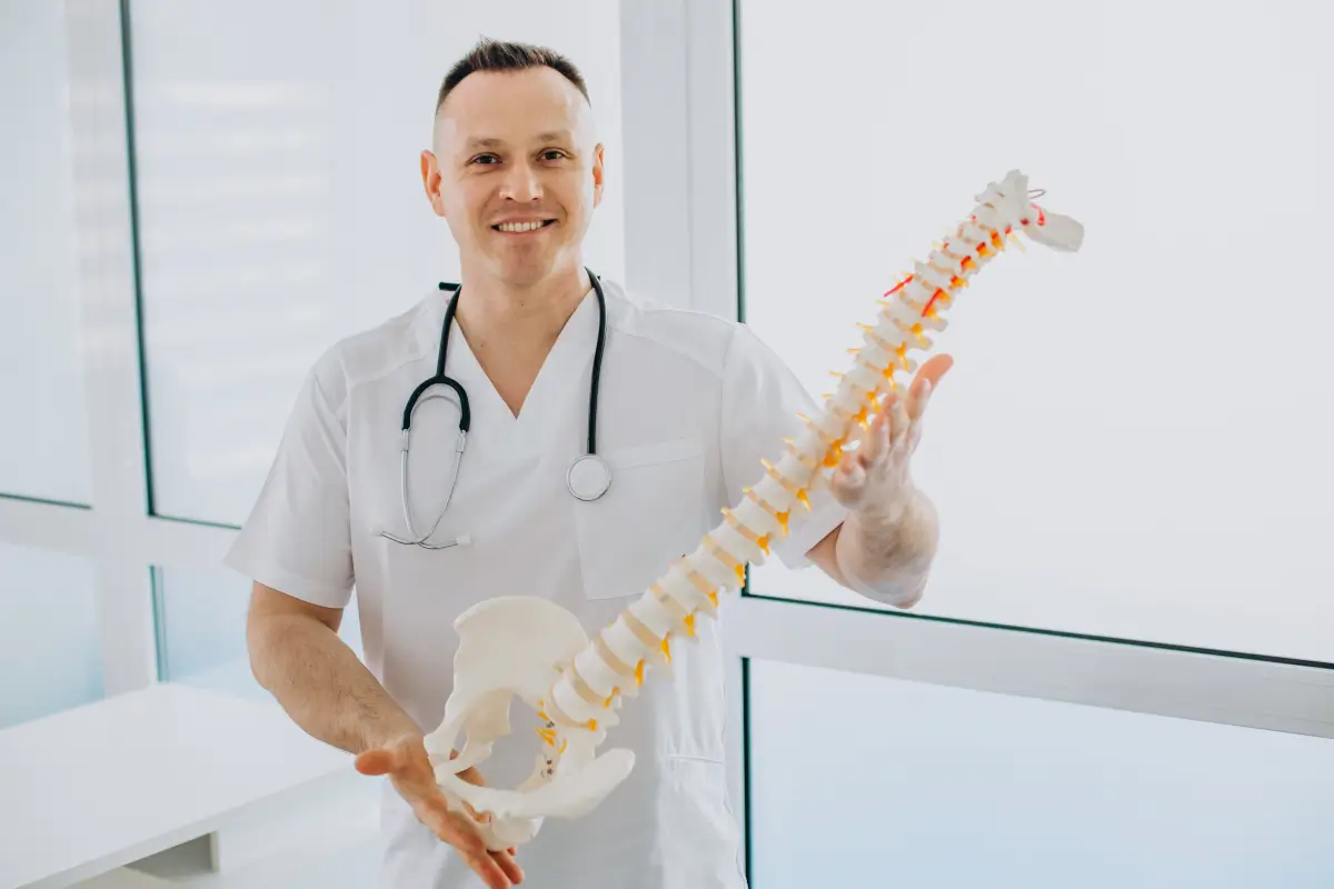 Ein lächelnder Physiotherapeut in weißer Arbeitskleidung zeigt ein anatomisches Modell der menschlichen Wirbelsäule, während er in einem hellen Behandlungsraum steht.