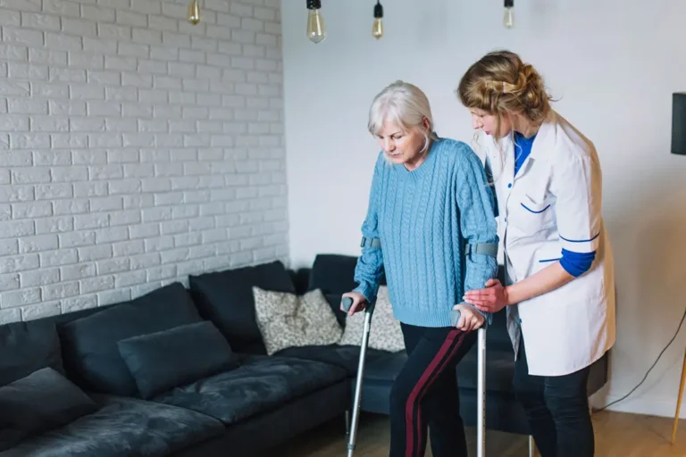Ältere Frau mit grauen Haaren und Krücken wird von einer Pflegerin in weißem Kittel beim Gehen unterstützt. Sie befinden sich in einem gemütlichen Wohnzimmer mit dunklem Sofa und weißer Backsteinwand.