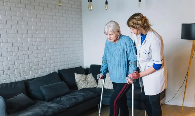 Ältere Frau mit grauen Haaren und Krücken wird von einer Pflegerin in weißem Kittel beim Gehen unterstützt. Sie befinden sich in einem gemütlichen Wohnzimmer mit dunklem Sofa und weißer Backsteinwand.