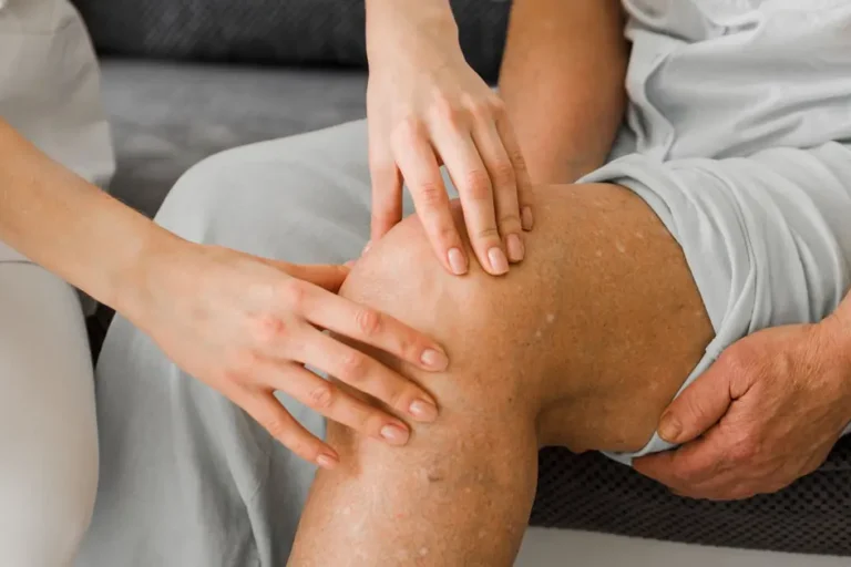 Nahaufnahme einer Physiotherapeutin, die das Knie eines älteren Patienten untersucht und sanft berührt.