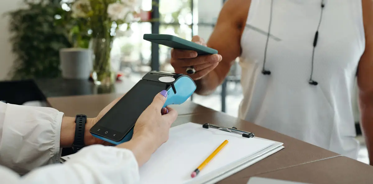 Person bezahlt kontaktlos mit einem Smartphone an einem Kartenlesegerät, während eine andere Person das Terminal hält. Szene in einem modernen, hellen Innenraum