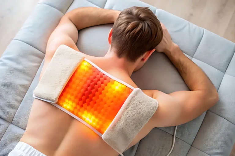 Mann liegt auf dem Bauch auf einem grauen Sofa und erhält eine Wärmetherapie mit einem beleuchteten Heizkissen auf dem Rücken. Die Anwendung dient zur Muskelentspannung und Schmerzlinderung.