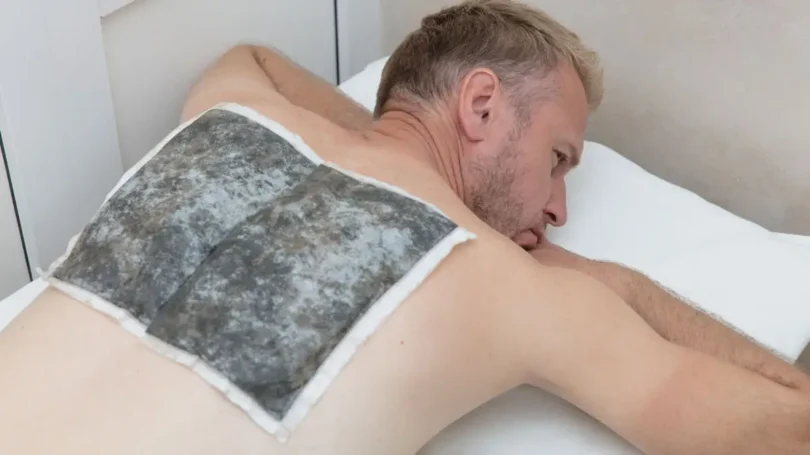 Ein Mann liegt entspannt auf einer Liege und erhält eine Fangopackung auf seinem Rücken während einer physiotherapeutischen Behandlung.