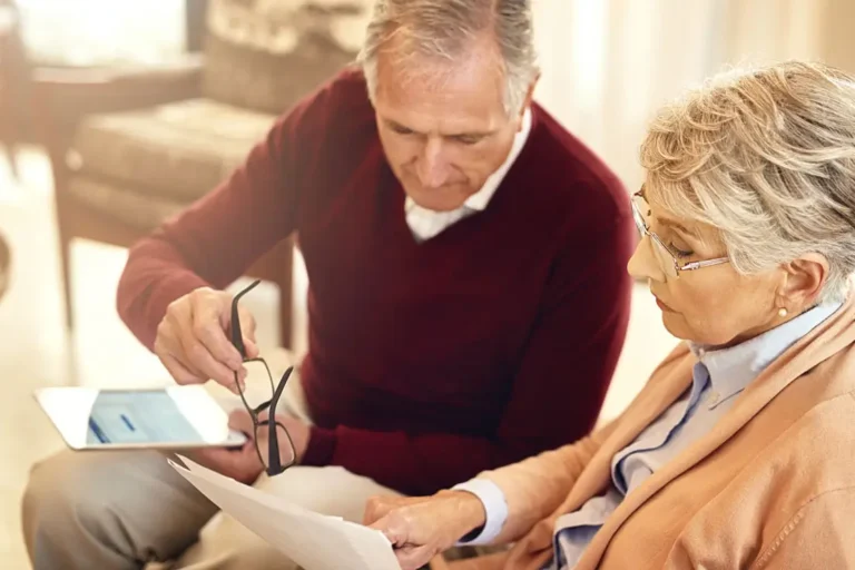 Älteres Paar sitzt gemeinsam auf einem Sofa und überprüft Dokumente. Der Mann hält eine Brille in der Hand, während die Frau konzentriert auf das Papier schaut. Im Hintergrund ist eine gemütliche Wohnzimmereinrichtung zu sehen.