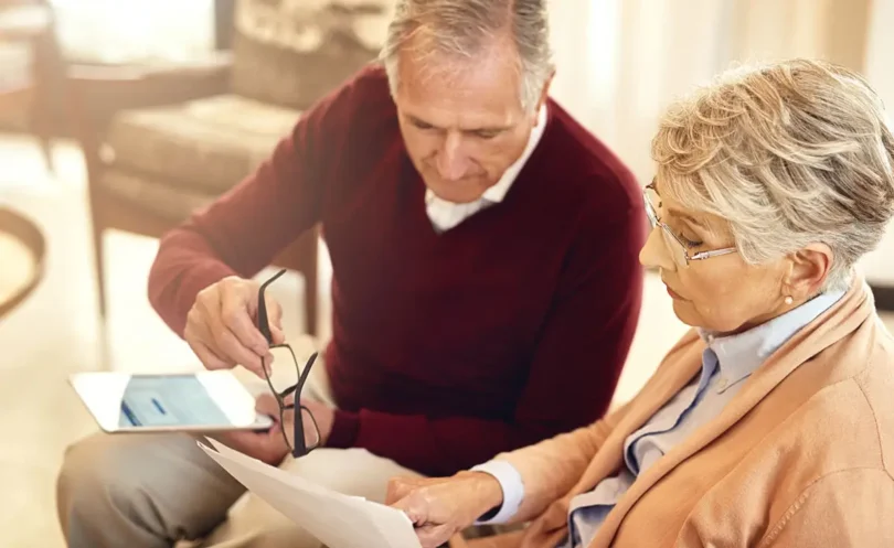 Älteres Paar sitzt gemeinsam auf einem Sofa und überprüft Dokumente. Der Mann hält eine Brille in der Hand, während die Frau konzentriert auf das Papier schaut. Im Hintergrund ist eine gemütliche Wohnzimmereinrichtung zu sehen.