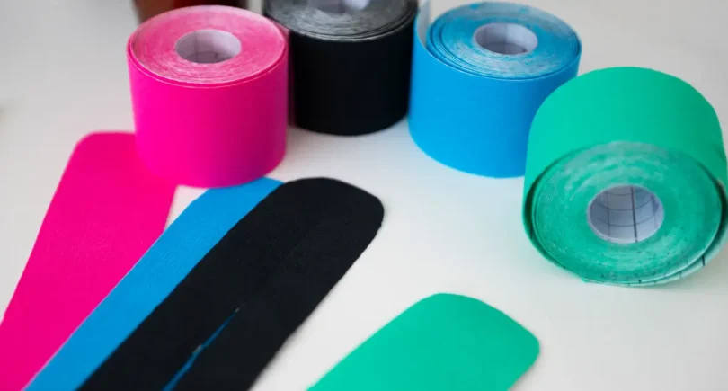 Mehrere bunte Kinesio-Tape-Rollen in Pink, Schwarz, Blau und Grün mit zugeschnittenen Tape-Streifen, die für physiotherapeutische Anwendungen und Rehabilitation genutzt werden.