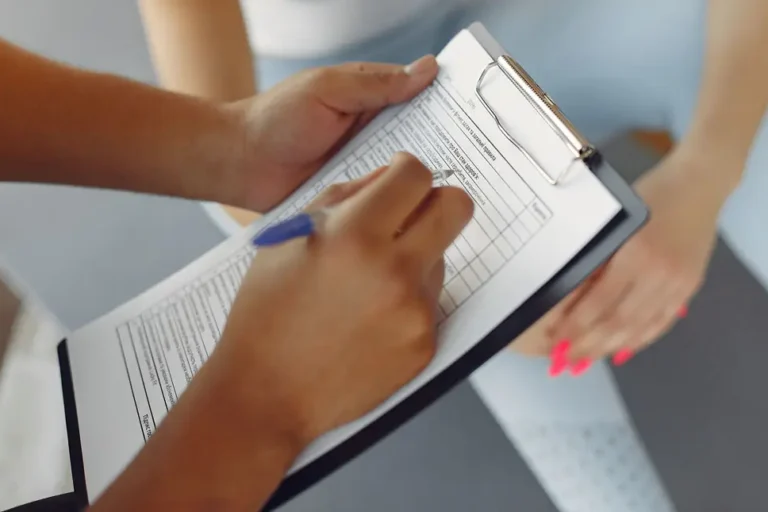 Nahaufnahme einer Person, die ein Formular auf einem Klemmbrett ausfüllt, während eine andere Person mit verschränkten Händen auf einer Gymnastikmatte sitzt.