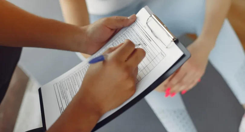 Nahaufnahme einer Person, die ein Formular auf einem Klemmbrett ausfüllt, während eine andere Person mit verschränkten Händen auf einer Gymnastikmatte sitzt.