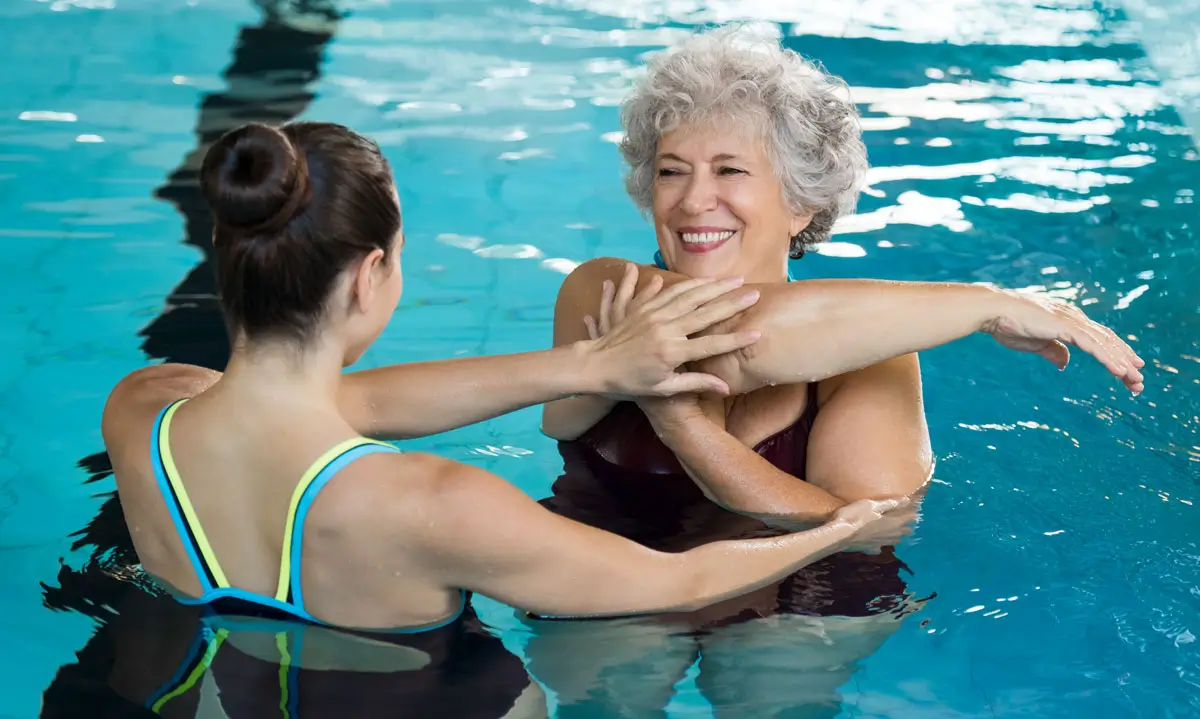 Eine ältere Frau nimmt an einer Wassertherapie teil, unterstützt von einer jungen Physiotherapeutin im Schwimmbecken.
