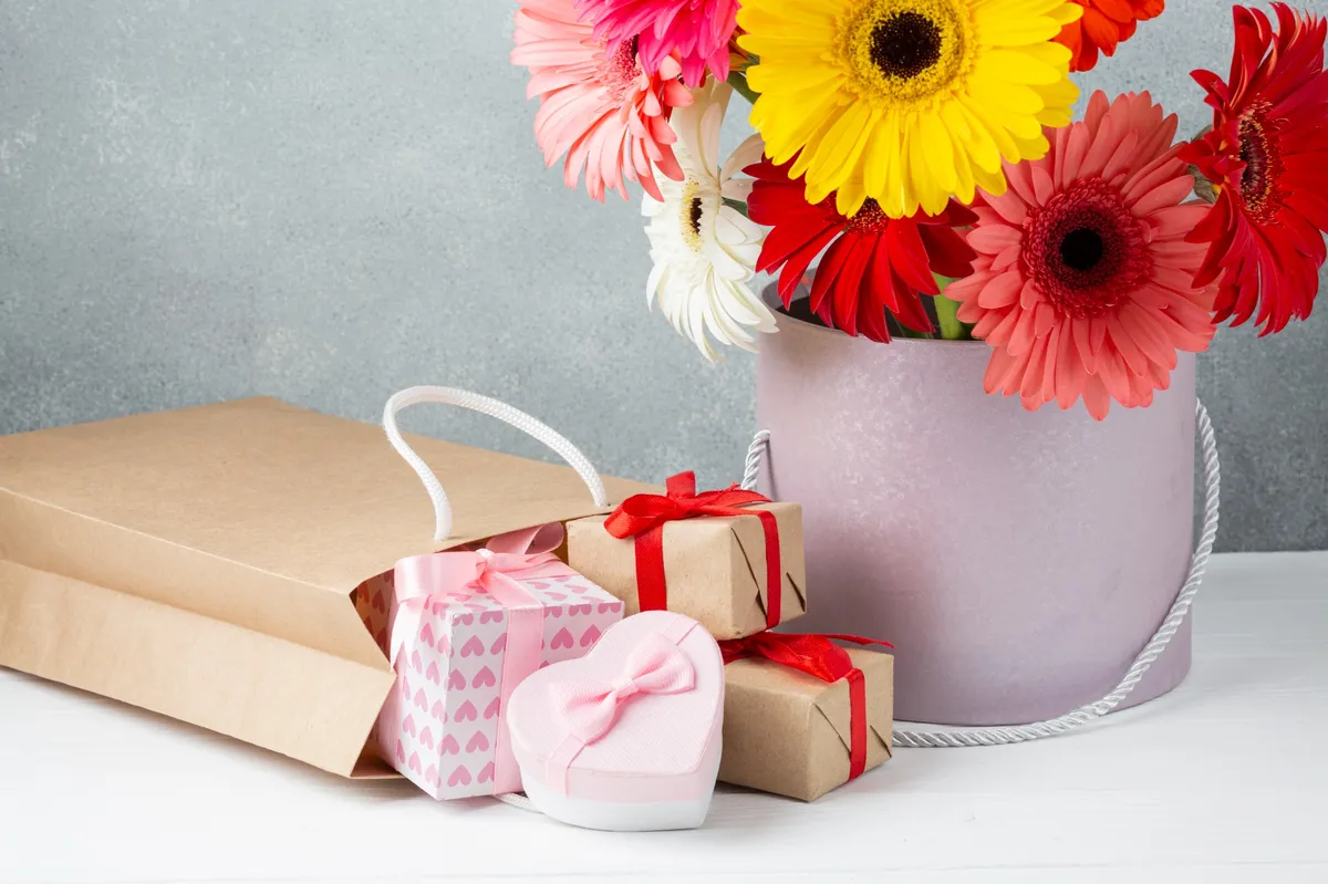 Bunte Gerbera-Blumen in einer Box mit mehreren Geschenken und einer Papiertüte auf weißem Tisch
