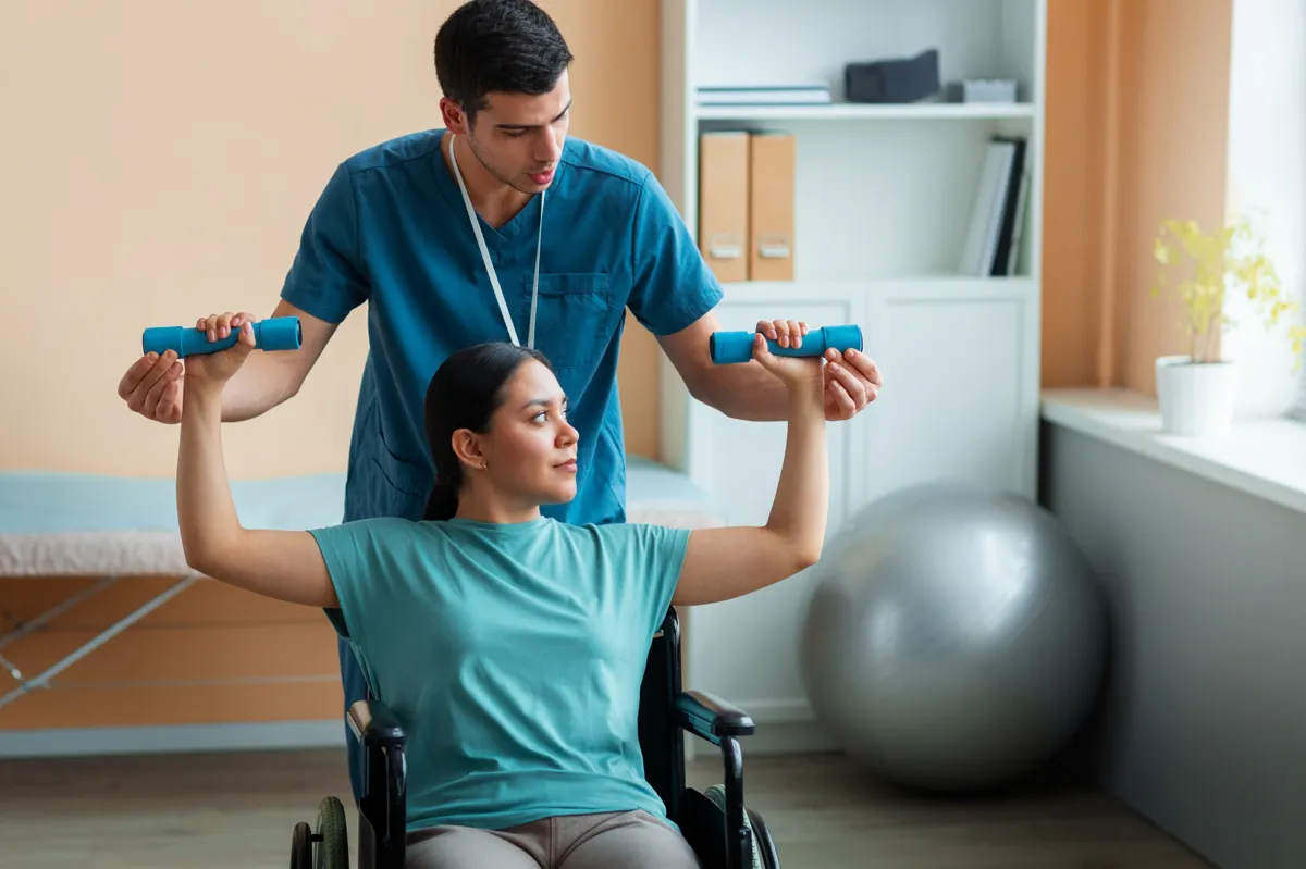 Physiotherapeut unterstützt eine Frau im Rollstuhl bei einer Rehabilitationsübung mit Hanteln in einer Klinik.