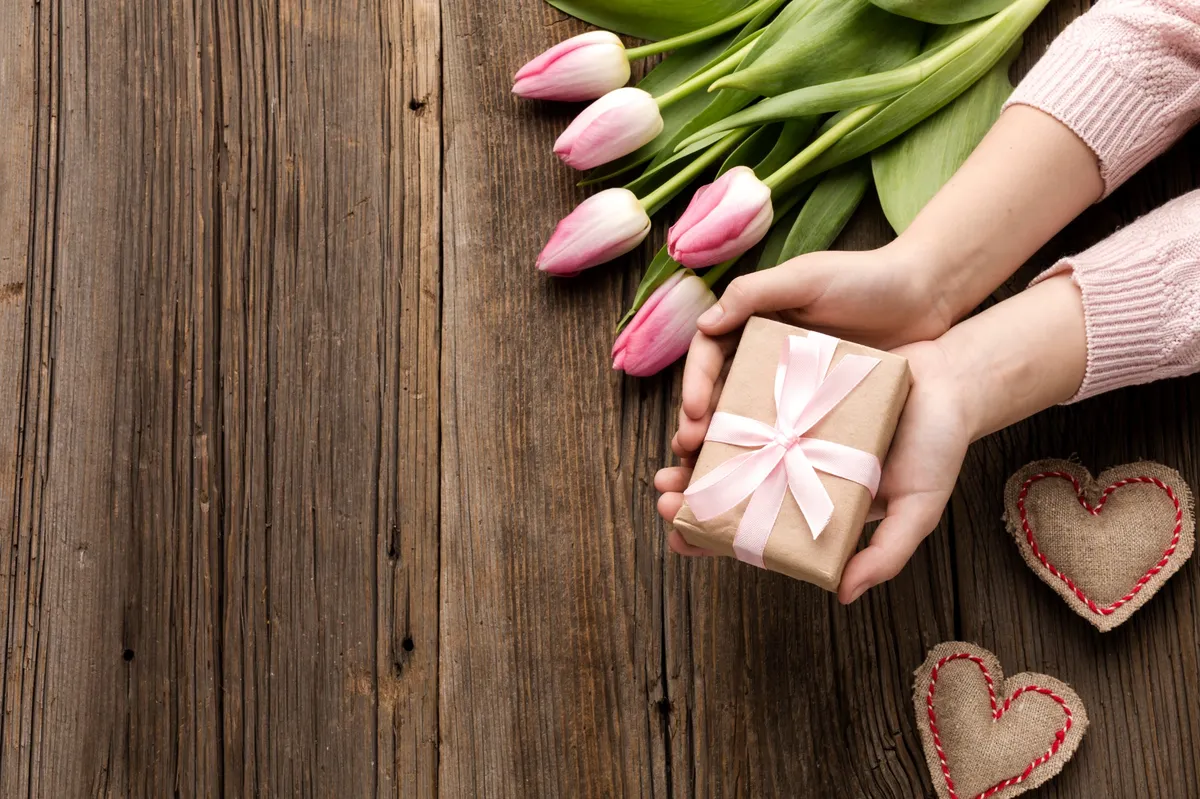 Hände halten ein Geschenk mit rosa Schleife neben rosa Tulpen auf einem Holztisch