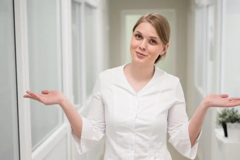 Junge Frau in medizinischer Kleidung steht in einem hellen Flur und zuckt fragend mit den Schultern – symbolisiert Unsicherheit oder Entscheidungsfindung im Gesundheitsbereich.