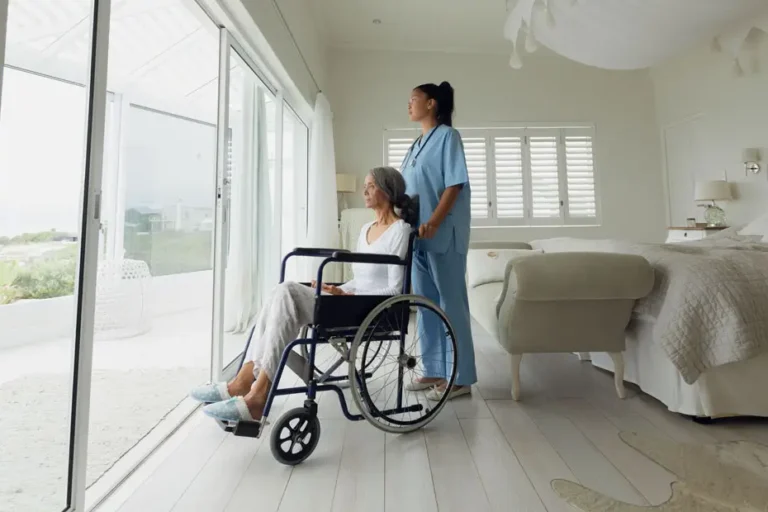 Pflegekraft steht neben einer älteren Frau im Rollstuhl in einem hellen, barrierefreien Einzelzimmer mit Blick auf eine Terrasse – Symbolbild für Ruhe und Privatsphäre in der Reha.