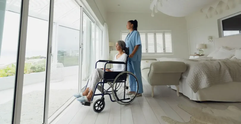 Pflegekraft steht neben einer älteren Frau im Rollstuhl in einem hellen, barrierefreien Einzelzimmer mit Blick auf eine Terrasse – Symbolbild für Ruhe und Privatsphäre in der Reha.