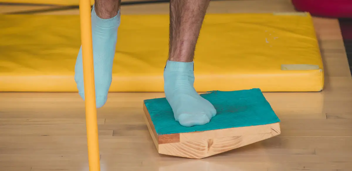 Person balanciert auf einem Bein mit blauen Socken auf einem schräg geneigten Holz-Wackelbrett, neben einer gelben Matte und Haltestange