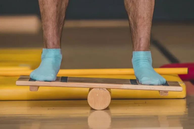 Person balanciert mit blauen Socken auf einem Holz-Wackelbrett in einer Therapiesituation, Fokus auf den Füßen und das Trainingsgerät