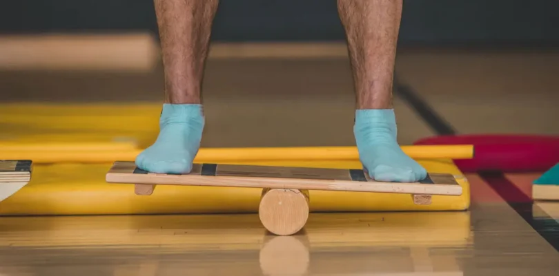 Person balanciert mit blauen Socken auf einem Holz-Wackelbrett in einer Therapiesituation, Fokus auf den Füßen und das Trainingsgerät