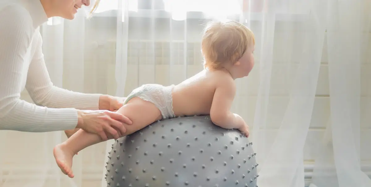 Baby liegt bäuchlings auf einem großen Therapieball, während eine Frau es behutsam hält – helle, freundliche Umgebung.