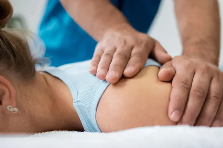 Physiotherapeut behandelt die Schulter einer Patientin in Bauchlage mit beiden Händen – Nahaufnahme in heller Praxisumgebung.