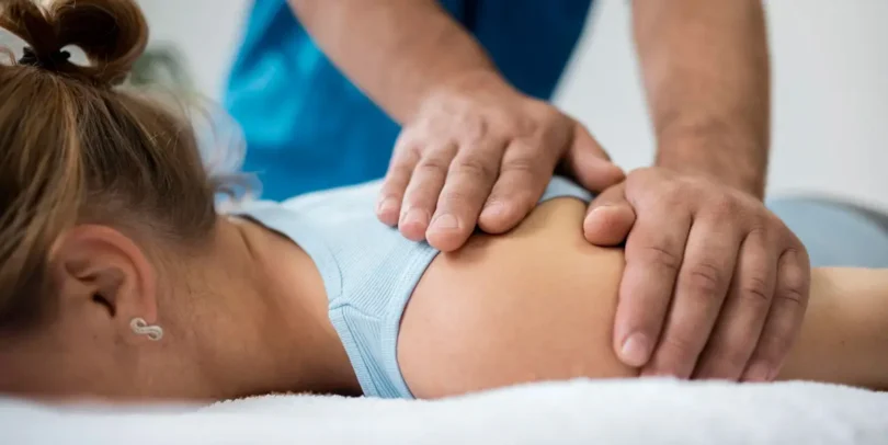 Physiotherapeut behandelt die Schulter einer Patientin in Bauchlage mit beiden Händen – Nahaufnahme in heller Praxisumgebung.