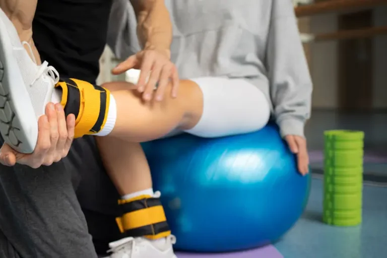 Physiotherapeut unterstützt Patientin bei einer Reha-Übung mit Gymnastikball und Fußmanschetten im Therapieraum.