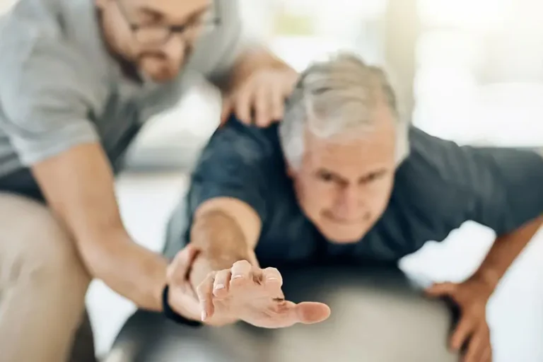Physiotherapeut unterstützt älteren Mann bei einer Gleichgewichts- und Dehnübung auf einem Gymnastikball zur Verbesserung der Mobilität und Rehabilitation.